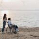 Slika prikazuje dvoje ljudi. Muškarac sjedi u kolicima dok je žena iza njega. Nalaze se na pješčanoj plaži uz more.
