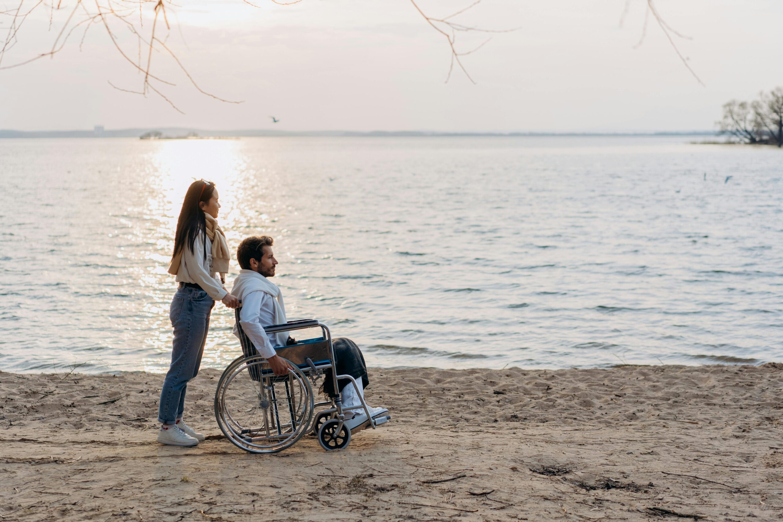 Slika prikazuje dvoje ljudi. Muškarac sjedi u kolicima dok je žena iza njega. Nalaze se na pješčanoj plaži uz more.