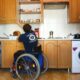Na slici je osoba u invalidskim kolicima koja pokušava dohvatiti čašu sa smeđeg visećeg ormara.
