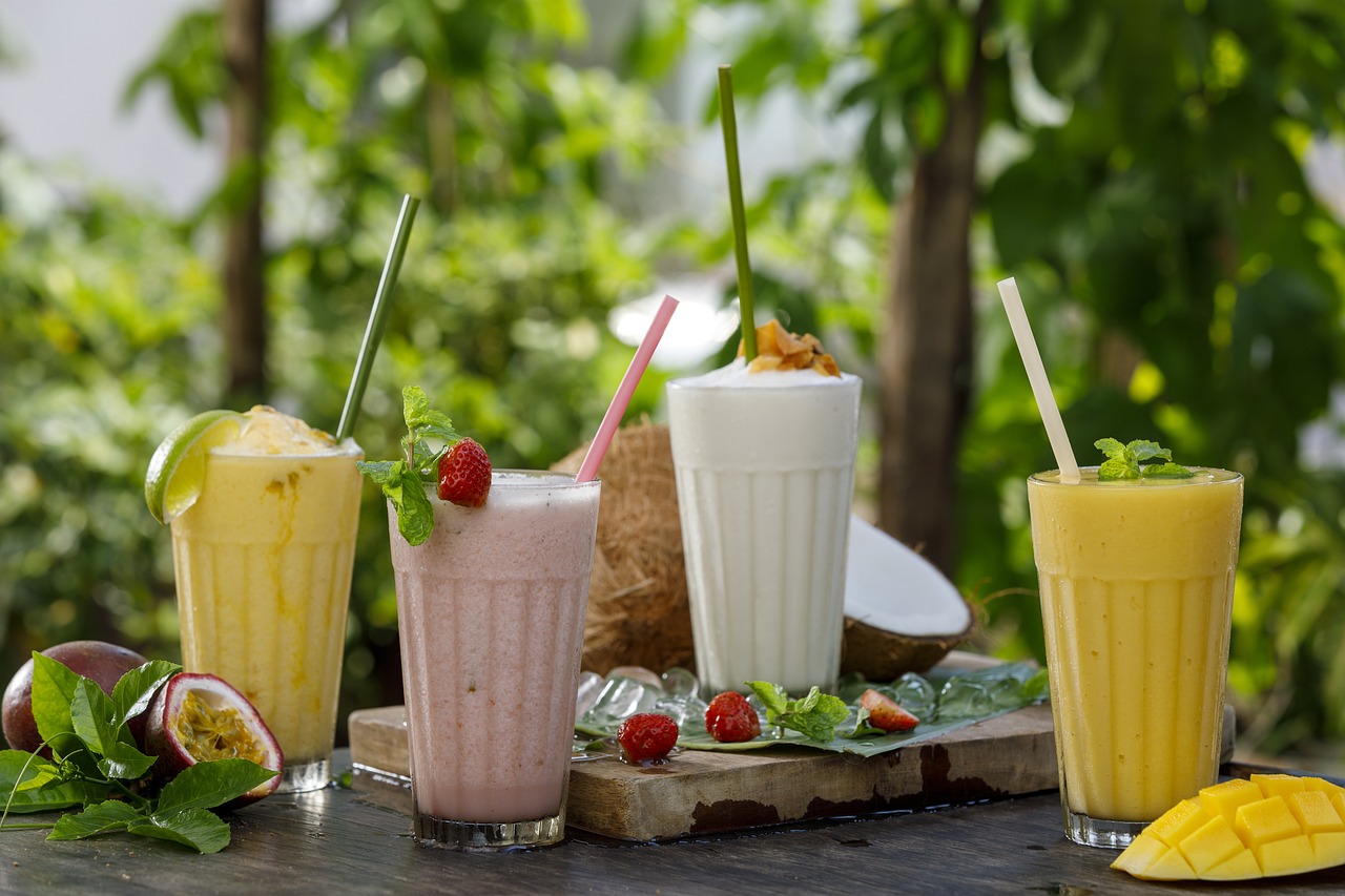 Na fotografiji su prikazana četiri različita napitka u visokim staklenim čašama s plastičnim slamkama, postavljeni na drvenu podlogu. S lijeva na desno, prvi napitak je žute boje s kriškom limuna i listićem mente, drugi je roze boje s jagodom na rubu čaše, treći je bijele boje u čaši koja nalikuje kokosovom orahu i ima komadiće voća na vrhu, a četvrti je također žute boje s listićem mente. U pozadini se nazire zelenilo biljaka. Fotografija bi mogla biti zanimljiva zbog prikaza raznolikih osvježavajućih ljetnih pića koja izgledaju privlačno i ukusno.