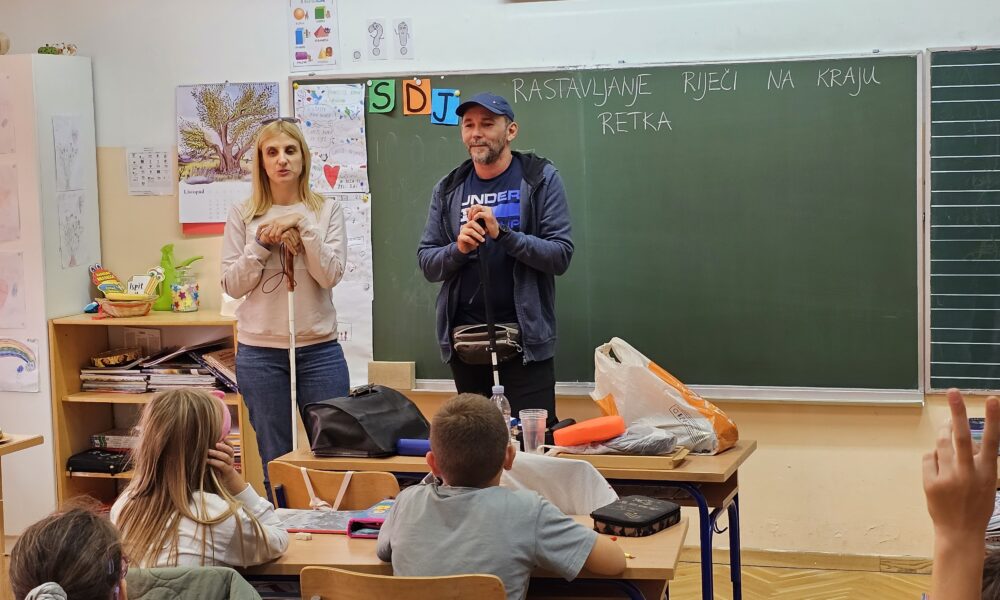 Na fotografiji je prikazana učionica u kojoj se nalaze dvoje odraslih i nekoliko djece. Odrasli stoje ispred ploče, dok djeca sjede za školskim klupama. Atmosfera izgleda mirno i edukativno.