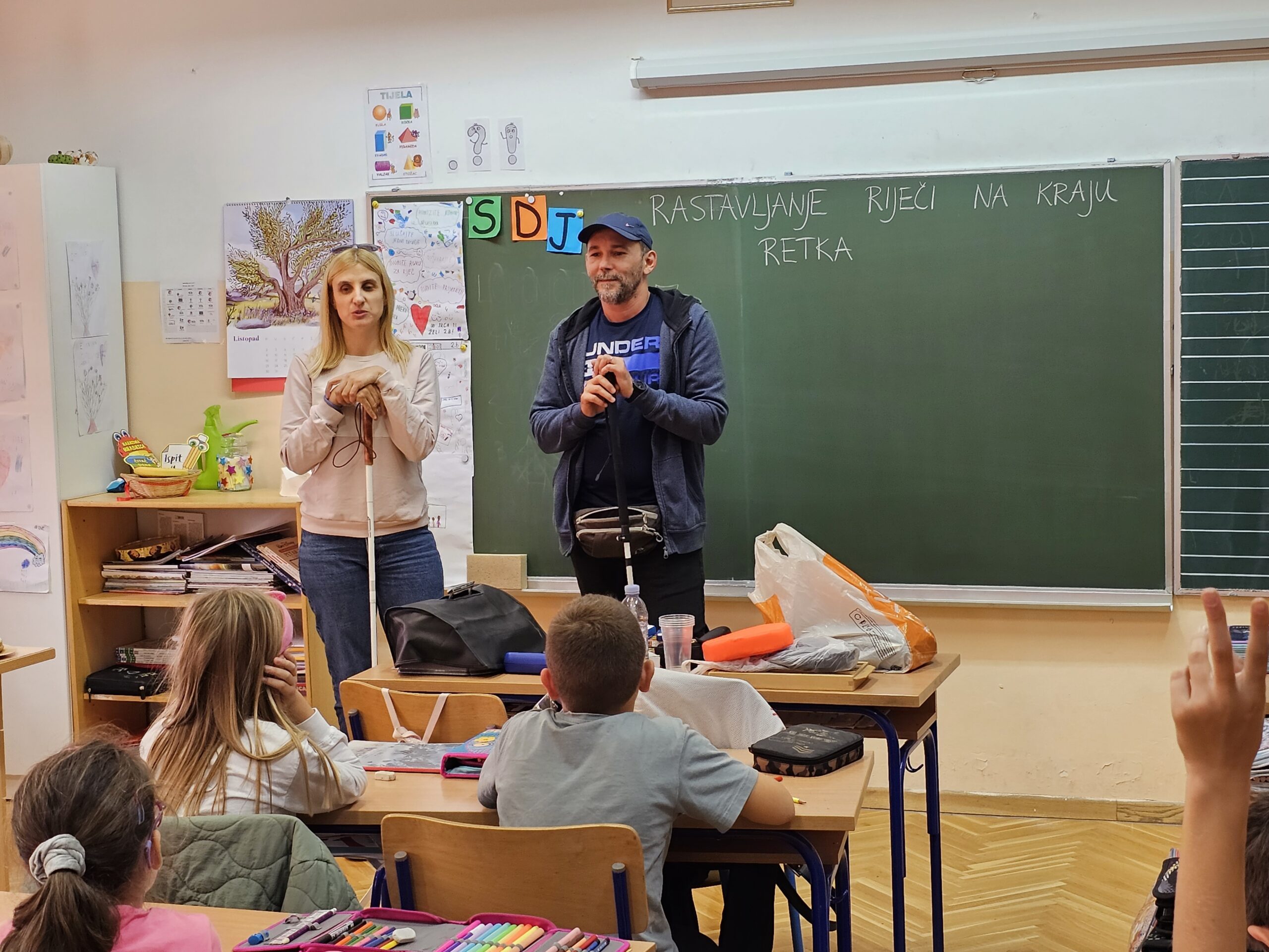 Na fotografiji je prikazana učionica u kojoj se nalaze dvoje odraslih i nekoliko djece. Odrasli stoje ispred ploče, dok djeca sjede za školskim klupama. Atmosfera izgleda mirno i edukativno.