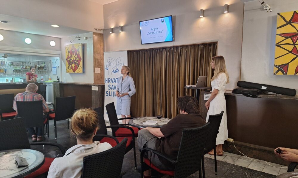 Fotografija prikazuje unutrašnjost prostorije u kojoj se održava konferencija. U prostoriji se nalazi nekoliko stolova s ljudima koji sjede okrenuti prema osobi koja stoji ispred televizora na kojem je prikazana prezentacija. Na zidu je vidljiva umjetnička slika apstraktnog dizajna. Televizor prikazuje matematički problem, ali tekst nije dovoljno jasan za transkripciju. Atmosfera na fotografiji djeluje opušteno, a osobe su usmjerene prema osobi koja prezentira i sadržaju na televizoru.