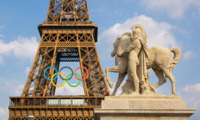 Na slici je prikazana Eiffelov toranj u Parizu, s velikim olimpijskim krugovima obješenim na sredini tornja. U prvom planu, desno, nalazi se kip muškarca s rimskom kacigom koji drži konja. Nebo je plavo s nekoliko bijelih oblaka. Kip i toranj simboliziraju spoj povijesti i modernih olimpijskih igara.