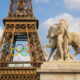 Na slici je prikazana Eiffelov toranj u Parizu, s velikim olimpijskim krugovima obješenim na sredini tornja. U prvom planu, desno, nalazi se kip muškarca s rimskom kacigom koji drži konja. Nebo je plavo s nekoliko bijelih oblaka. Kip i toranj simboliziraju spoj povijesti i modernih olimpijskih igara.