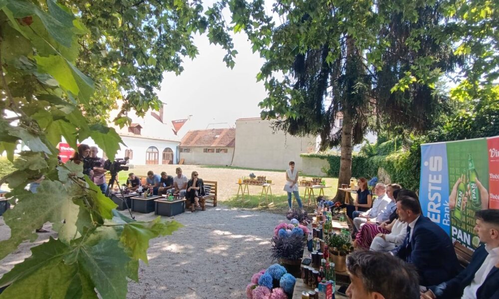 Skupina ljudi sjedi vani u hladu drveća, okupljeni oko stolova s pićem i cvijećem. U pozadini se vidi bijela zgrada s crvenim krovom. Ispred okupljenih stoji osoba koja drži govor. Atmosfera je opuštena, a događaj izgleda kao neka vrsta press konferencije ili društvenog okupljanja na otvorenom.