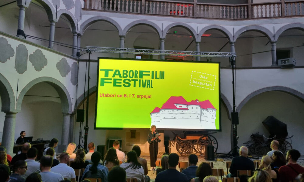 Na slici se nalazi unutrašnjost povijesnog zdanja s visokim lukovima i arkadama. U sredini slike je velika projekcijska platna s jarko žutim pozadinom na kojoj piše "TABOR FILM FESTIVAL" u zelenoj boji, te crveni tekst ispod "Utabori se 6. i 7. srpnja!" Desno od ovog teksta nalazi se ilustracija dvorca i obavijest "Ulaz na sve programe je besplatan" u bijeloj boji unutar okvira isprekidane linije. Ispred platna sjedi publika koja gleda prezentaciju, a ispred njih stoji govornik. U prostoriji se također nalaze razni stari predmeti poput kola i drugih povijesnih artefakata.