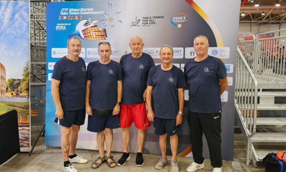 Na slici je grupa od pet starijih muškaraca, svi obučeni u tamnoplave majice i sportske hlače, stojeći ispred pozadinskog panoa na kojem piše "ITTF World Masters Table Tennis Championships Rome 2024". Svi su nasmijani i stoje jedno pored drugog u sportskom objektu.