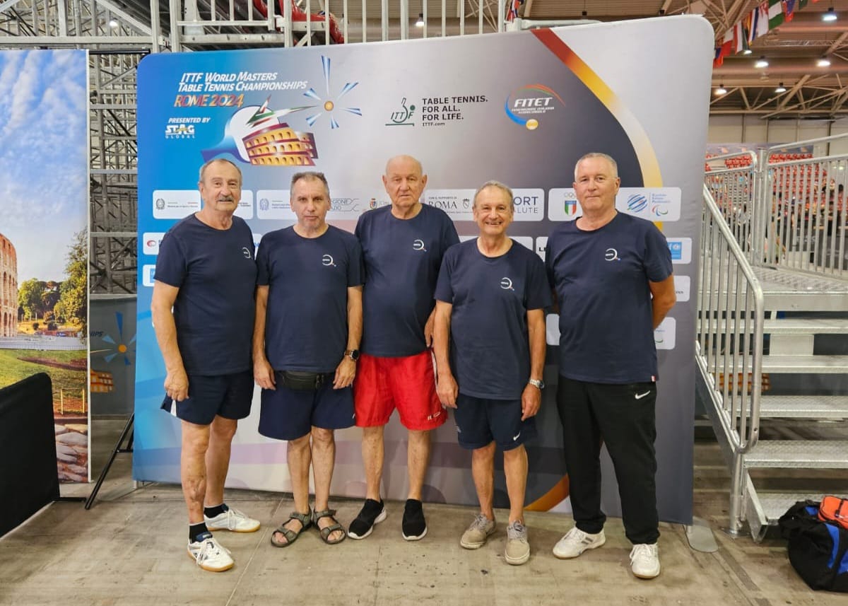 Na slici je grupa od pet starijih muškaraca, svi obučeni u tamnoplave majice i sportske hlače, stojeći ispred pozadinskog panoa na kojem piše "ITTF World Masters Table Tennis Championships Rome 2024". Svi su nasmijani i stoje jedno pored drugog u sportskom objektu.