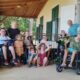 Na slici je prikazana grupa djece i odraslih ispred kuće s drvenim trijemom. Neki od njih su u invalidskim kolicima, a svi su nasmijani i izgledaju sretno. Okupljeni su u opuštenoj atmosferi, kao na obiteljskom okupljanju.