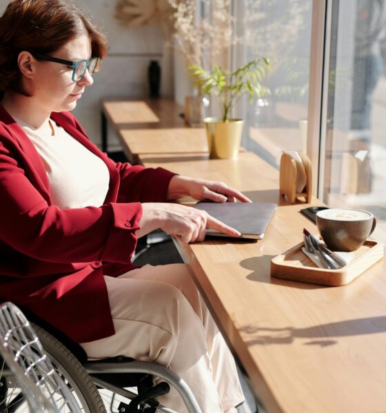 Na slici je osoba u invalidskim kolicima koja se priprema otvoriti laptop. Ispred nje je prozor, a na smeđem stolu uz laptop nalazi se i kava, pribor za jelo i dvije vaze cvijeća. Osoba nosi naočale, bijelu majicu i crveni pulover.