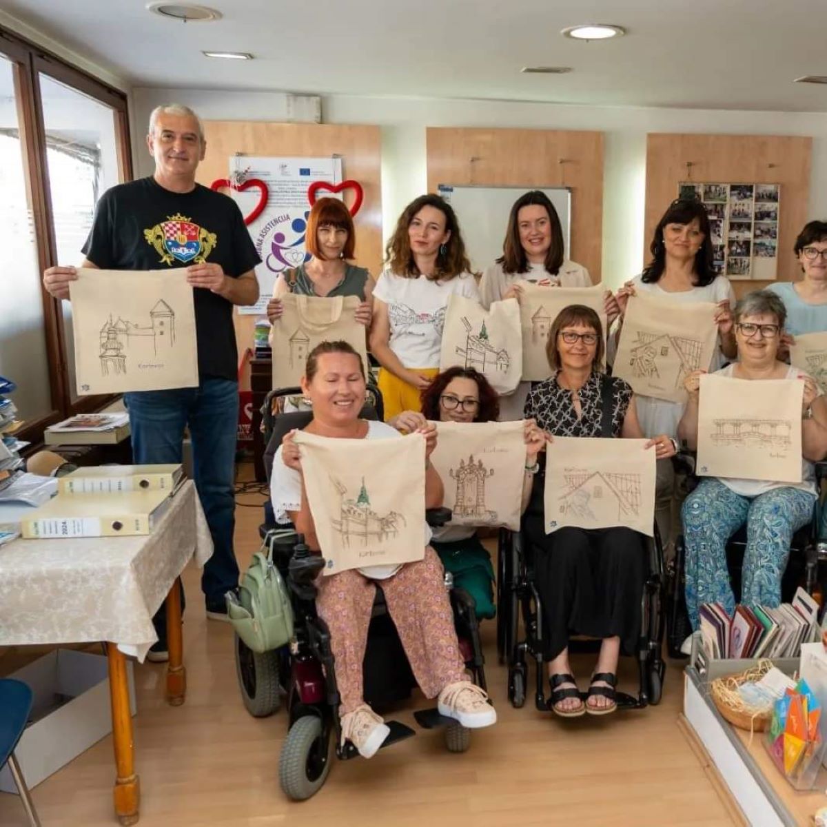 Na fotografiji je grupa ljudi koji drže papire s crtežima. Nalaze se u prostoriji s drvenim namještajem, a neki od njih sjede u invalidskim kolicima. Svaka vrećica prikazuje građevinu koja podsjeća na crkvu ili katedralu. Izgleda da su sudjelovali u umjetničkoj radionici ili aktivnosti