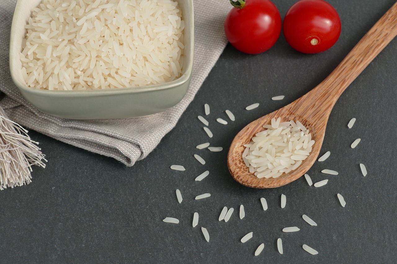 Na fotografiji je prikazana zdjela puna bijele riže postavljene na sivu podlogu uz koju se nalazi drvena žlica također napunjena rižom. Pored zdjele i žlice su dvije crvene rajčice koje pružaju kontrast boja u odnosu na bijelu rižu i sivu podlogu. Ova fotografija može biti zanimljiva ili relevantna zbog prikaza hrane u estetski ugodnom aranžmanu koji naglašava teksture i boje, što je često važno u kulinarskoj fotografiji.