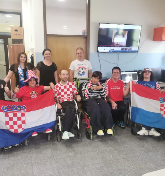 Na fotografiji je grupa od osam osoba u unutrašnjem prostoru. Neki od njih drže zastavu Hrvatske, koja je crveno-bijelo-plava s grbom u sredini. Neke osobe su u invalidskim kolicima. U pozadini se vidi televizor na zidu i vrata s natpisom. Lica osoba su zamagljena radi zaštite privatnosti.
