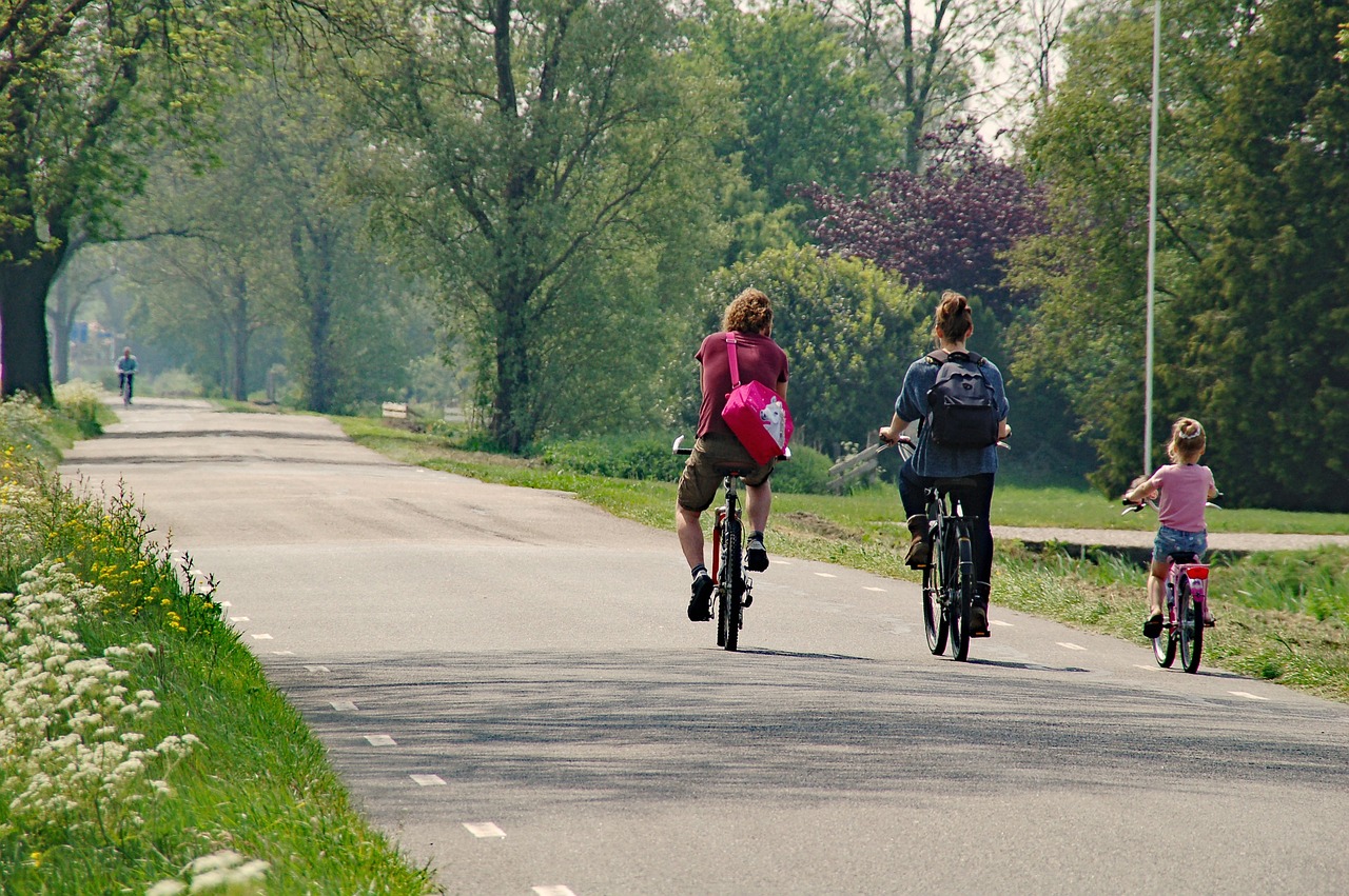Fotografija prikazuje troje ljudi koji voze bicikle na asfaltiranom putu. Osoba s lijeve strane nosi ruksak ružičaste boje, dok osoba u sredini ima tamnu odjeću. Treća osoba, koja je dijete, vozi manji bicikl i nalazi se desno. Okruženje je prirodno i mirno, sa zelenilom i drvećem uz put te svijetlim nebom u pozadini. Ova slika može biti zanimljiva ili relevantna jer prikazuje obiteljsku rekreaciju na otvorenom te promiče zdrav životni stil.