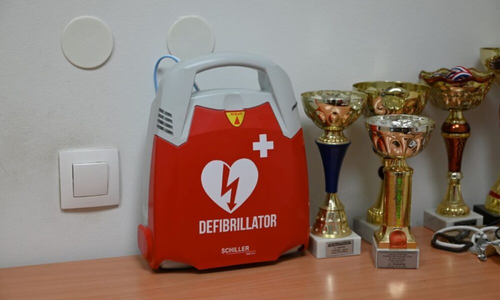 Na fotografiji je crveni defibrilator marke Schiller postavljen na stolu. U pozadini se vide sportski trofeji različitih veličina i oblika, što ukazuje na to da bi lokacija mogla biti sportska ustanova ili klub. Defibrilator je medicinski uređaj koji se koristi za spašavanje života u slučaju srčanog zastoja, što ga čini značajnim i relevantnim predmetom na slici. Na zidu iznad stola nalaze se dva bijela okrugla objekta koji izgledaju kao dio nekog sustava, možda alarma ili termostata, te jedan kvadratni prekidač.