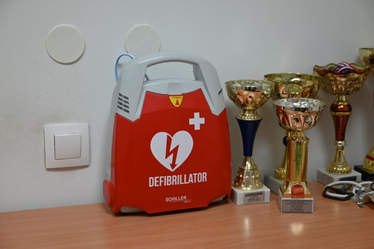 Na fotografiji je crveni defibrilator marke Schiller postavljen na stolu. U pozadini se vide sportski trofeji različitih veličina i oblika, što ukazuje na to da bi lokacija mogla biti sportska ustanova ili klub. Defibrilator je medicinski uređaj koji se koristi za spašavanje života u slučaju srčanog zastoja, što ga čini značajnim i relevantnim predmetom na slici. Na zidu iznad stola nalaze se dva bijela okrugla objekta koji izgledaju kao dio nekog sustava, možda alarma ili termostata, te jedan kvadratni prekidač.