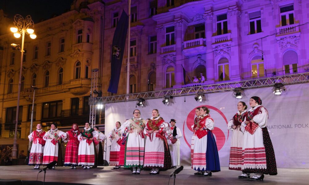 Na slici je prikazana folklorna izvedba na otvorenom, s grupom plesača u tradicionalnim narodnim nošnjama na pozornici. Pozadina je osvijetljena ljubičastim svjetlom, a iza njih se vidi fasada stare zgrade. Publika je okupljena ispred pozornice.