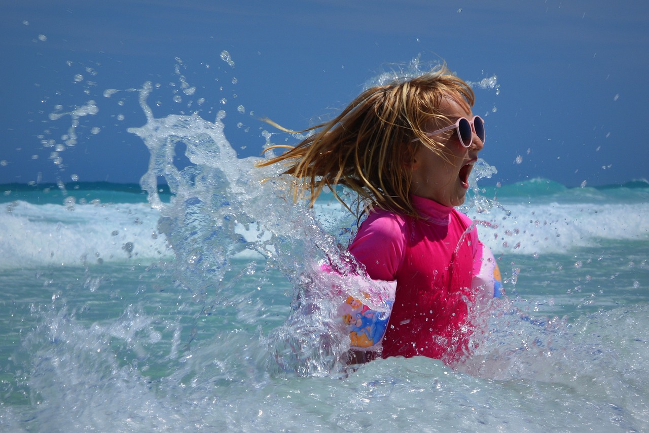 Fotografija prikazuje djevojčicu koja se nalazi u moru dok je okružena prskanjem vode. Osoba nosi ružičastu majicu, a lice joj je zamagljeno radi zaštite privatnosti. U pozadini se vidi plavo nebo i valovi na površini mora. Slika je dinamična i prikazuje interakciju osobe s valovima i vodom, stvarajući kontrast između mirnog neba i živahnog prskanja vode.