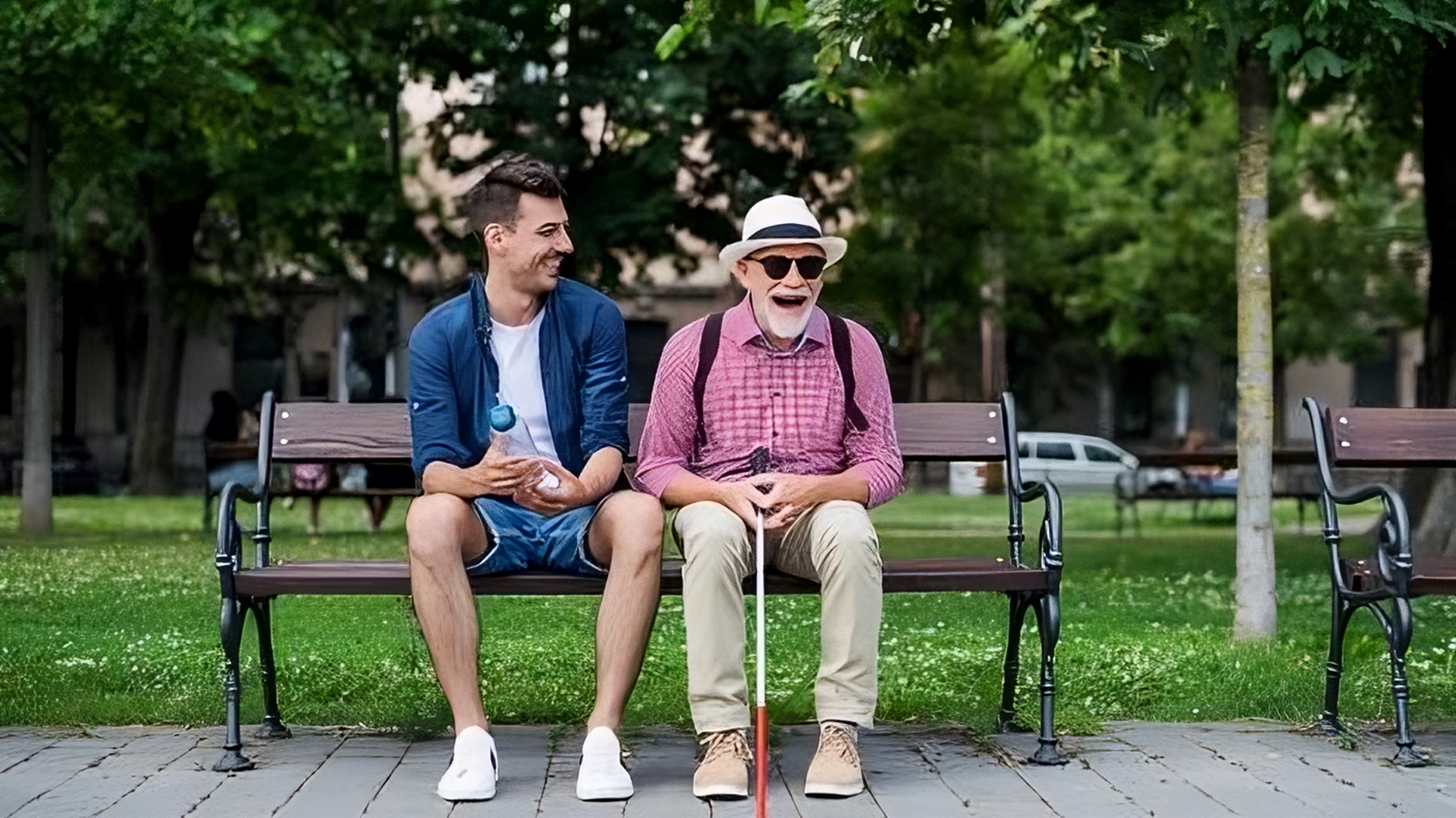 Na slici su dva muškarca koji sjede na klupi u parku. Lijevo je mlađi muškarac u ležernoj odjeći: plavoj jakni, bijeloj majici i kratkim hlačama, s osmijehom na licu dok gleda starijeg muškarca. Desno je stariji muškarac s bijelom bradom, koji nosi bijeli šešir, sunčane naočale, ružičastu kariranu košulju, bež hlače i ruksak. On također izgleda sretno i razgovara, držeći bijeli štap za slijepe. Oko njih je bujna zelena vegetacija s drvećem i travnjakom, a u pozadini se vidi parkirana vozila i zgrade.