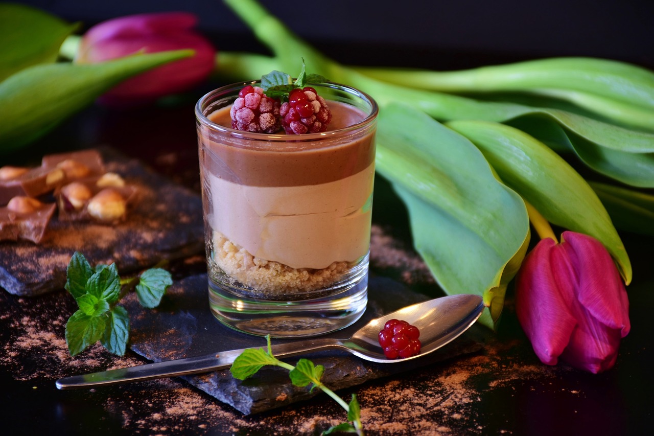 Na fotografiji je desert u staklenoj čaši koji se sastoji od slojeva, s najdonjim slojem koji izgleda kao mrvičasti biskvit, srednji sloj kremaste teksture svijetle boje i gornji sloj tamnije kremaste teksture. Desert je ukrašen malinama na vrhu. Uz čašu su raspoređeni listovi biljke, svježe maline, komadići čokolade i tulipan. Pozadina je tamna koja ističe boje deserta i dodataka. Desert izgleda ukusno i pažljivo pripremljen, što bi moglo biti zanimljivo za ljubitelje slastica ili za one koji traže inspiraciju za prezentaciju hrane.