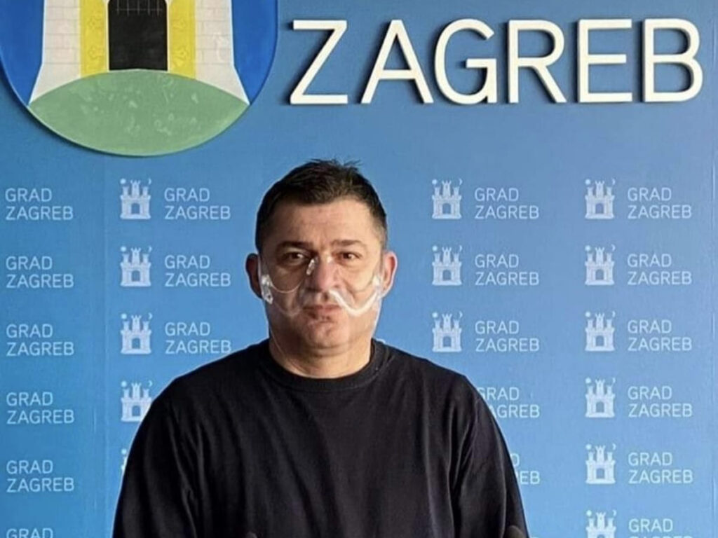 Muškarac tamne kose stoji ispred plavog zida s logom Grada Zagreba. Na licu nosi prozirnu zaštitnu masku, a odjeven je u crnu majicu.