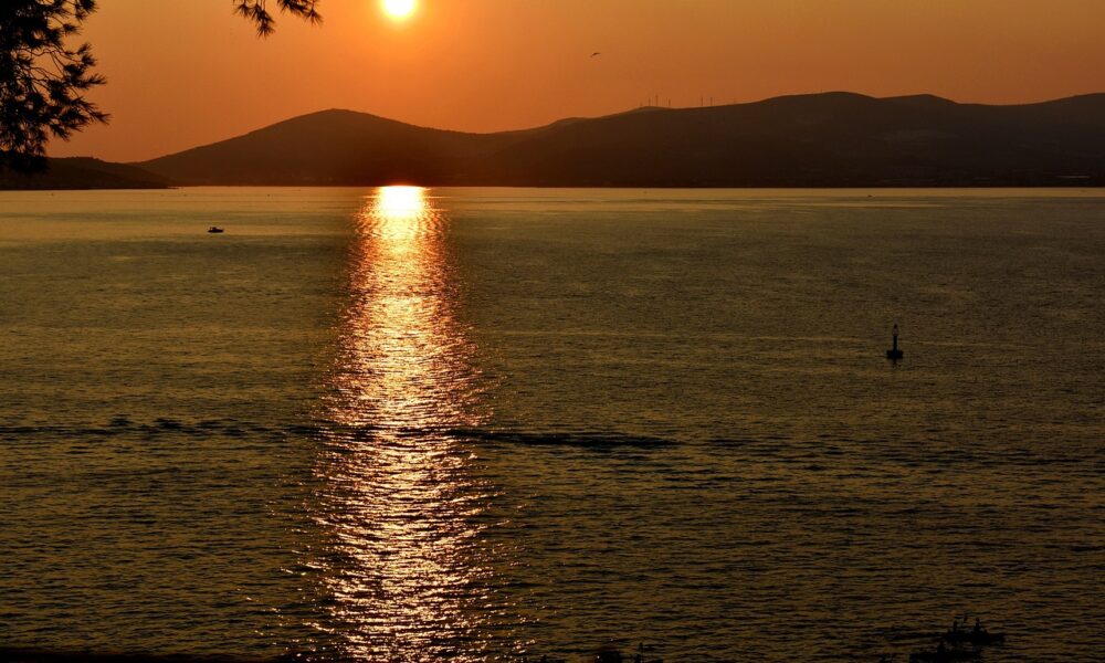 Na slici je prikazan zalazak sunca nad morem. Sunce je nisko na horizontu, reflektirajući svoje zlatne zrake na površini vode. U pozadini se vide tamne siluete brda.