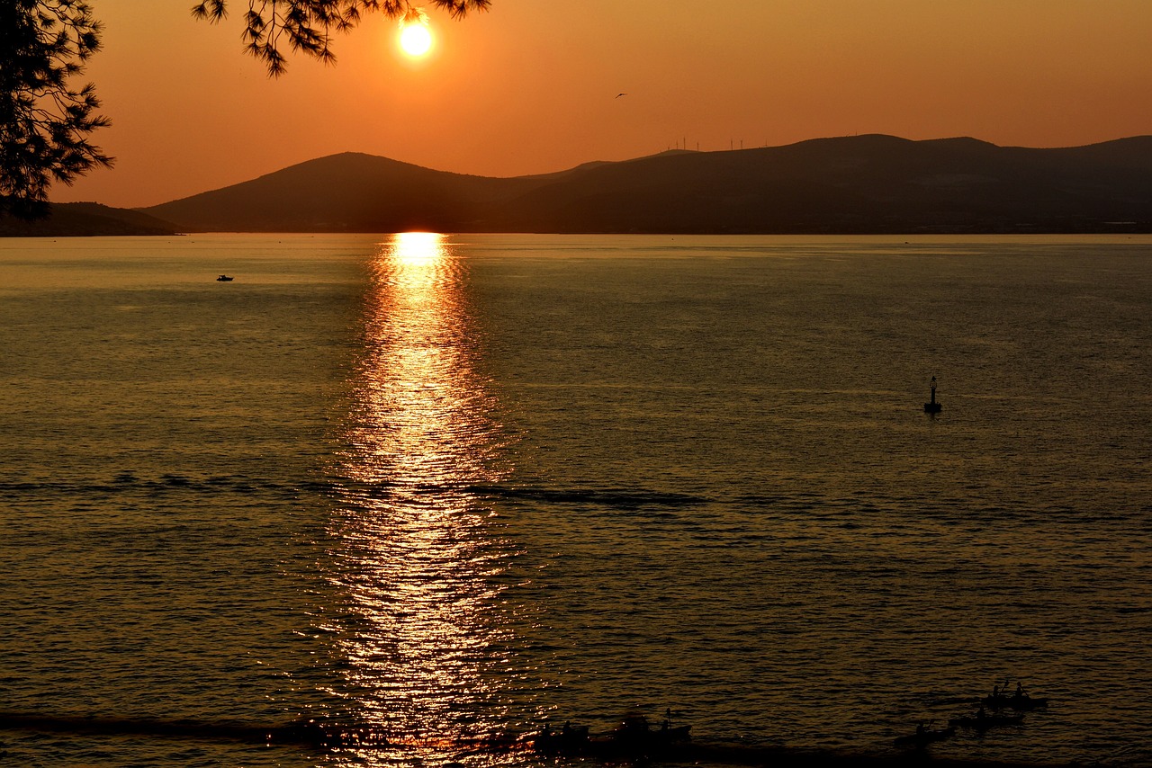 Na slici je prikazan zalazak sunca nad morem. Sunce je nisko na horizontu, reflektirajući svoje zlatne zrake na površini vode. U pozadini se vide tamne siluete brda.