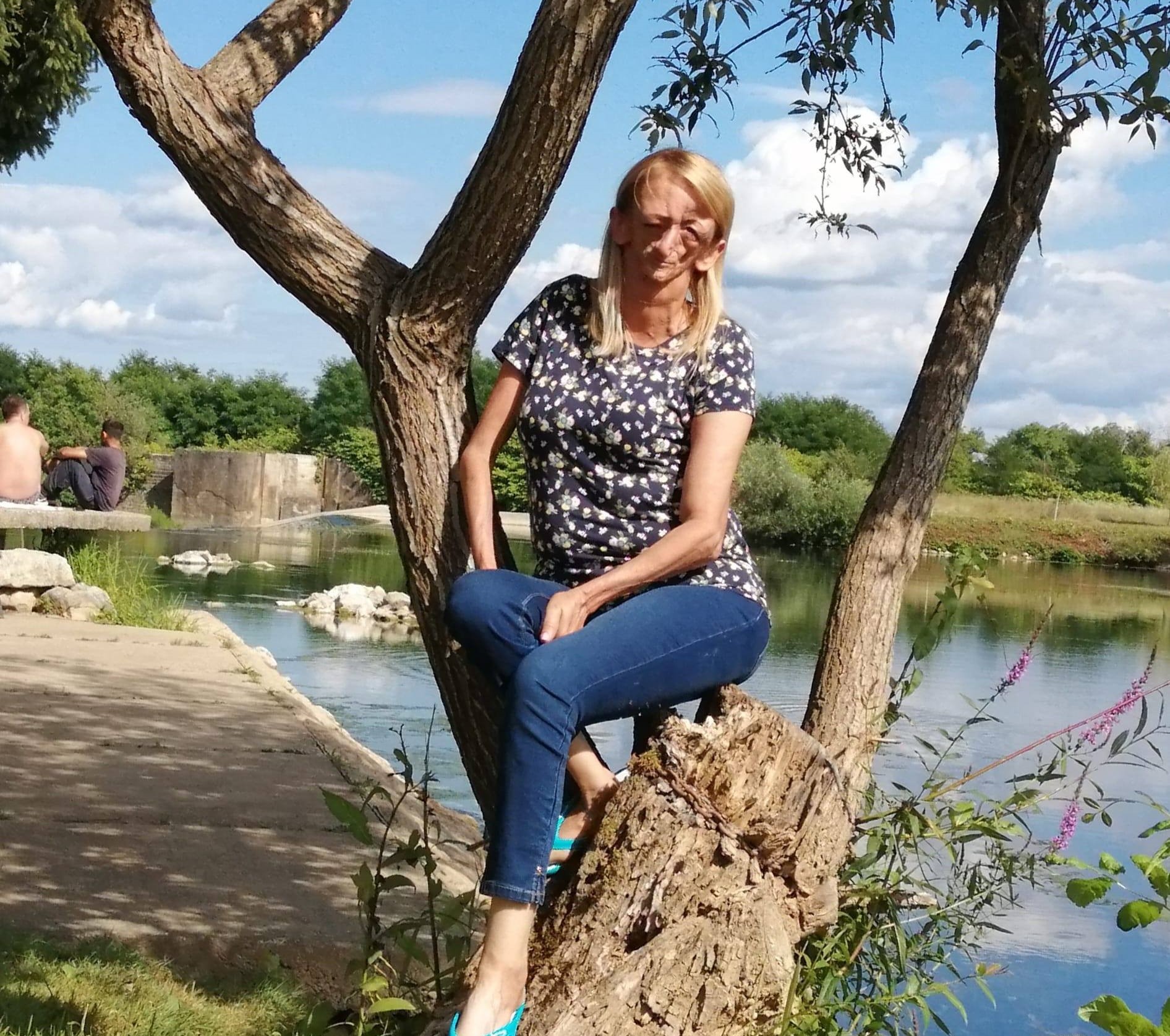 Na fotografiji je osoba koja sjedi na stablu pored jezera. Osoba nosi plave traperice i majicu s uzorkom, a lice joj je zamagljeno radi privatnosti. U pozadini se vide voda, zelenilo i drugi ljudi koji uživaju u prirodi. Ova fotografija može biti zanimljiva zbog opuštajuće atmosfere prirodnog okruženja.