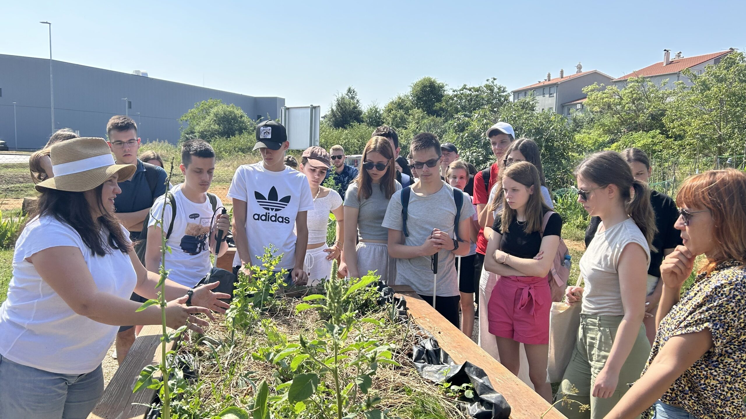 Na fotografiji je grupa ljudi koja stoji oko drvene ograde. Ljudi su različito odjeveni, neki nose šešire i sunčane naočale, a neki su bez njih. Izgleda kao da su u posjetu nekom vrtu ili parku jer se u pozadini vide zgrade i drveće.