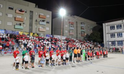 Na slici su nogometaši i sudci koji se nalaze na vanjskom sportskom terenu, a ispred njih nalaze se djeca. Publika je od crvenih i bijelih papirića napravila i podigla hrvatsku šahovnicu. Ovdje se vjerojatno radi o nogometnom turniru. Iznad publike nalaze se napisana imena sponzora natjecanja.