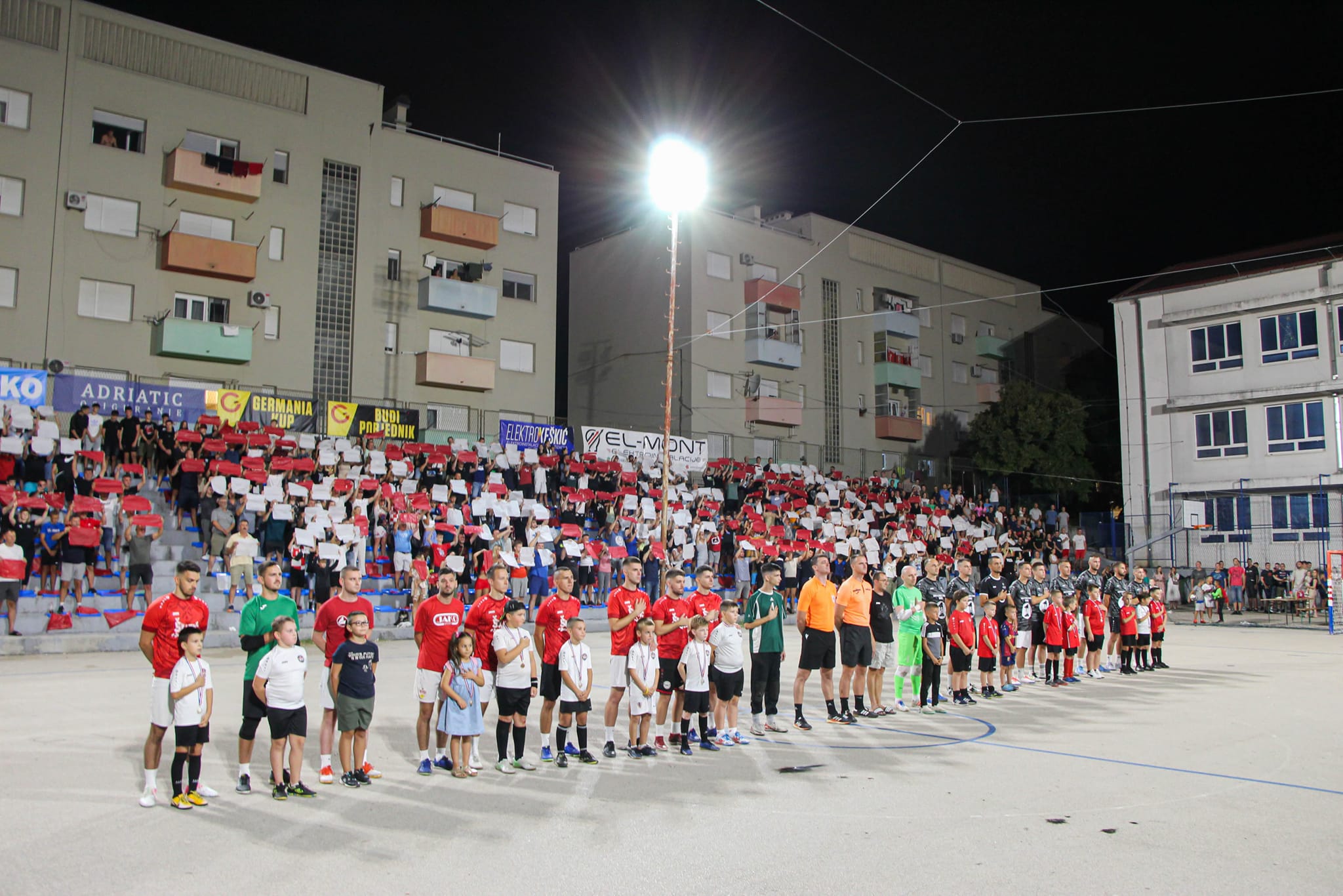 Na slici su nogometaši i sudci koji se nalaze na vanjskom sportskom terenu, a ispred njih nalaze se djeca. Publika je od crvenih i bijelih papirića napravila i podigla hrvatsku šahovnicu. Ovdje se vjerojatno radi o nogometnom turniru. Iznad publike nalaze se napisana imena sponzora natjecanja.