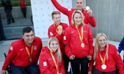 Na slici su sportaši s invaliditetom, Neki od njih sjede u kolicima, neki stoje, a jedna je osoba na štakama. Sportaši oko vrata nose osvojene medalje, a obučeni su u crvene trenirke na kojima piše 'Hrvatski parolimpijski odbor'.