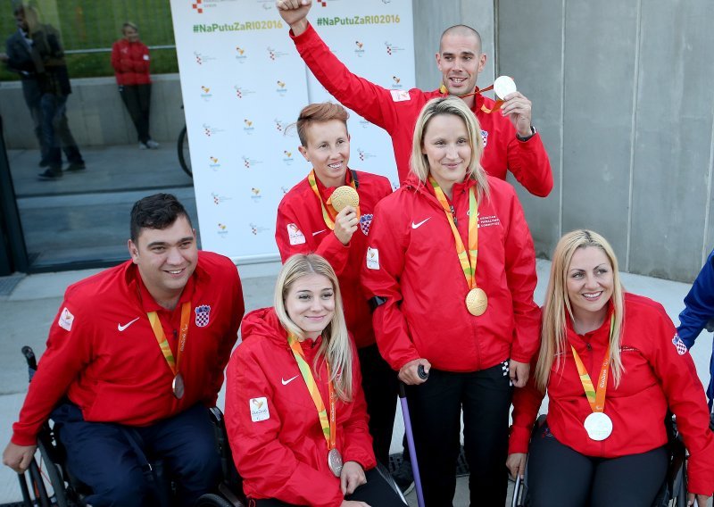 Na slici su sportaši s invaliditetom, Neki od njih sjede u kolicima, neki stoje, a jedna je osoba na štakama. Sportaši oko vrata nose osvojene medalje, a obučeni su u crvene trenirke na kojima piše 'Hrvatski parolimpijski odbor'.