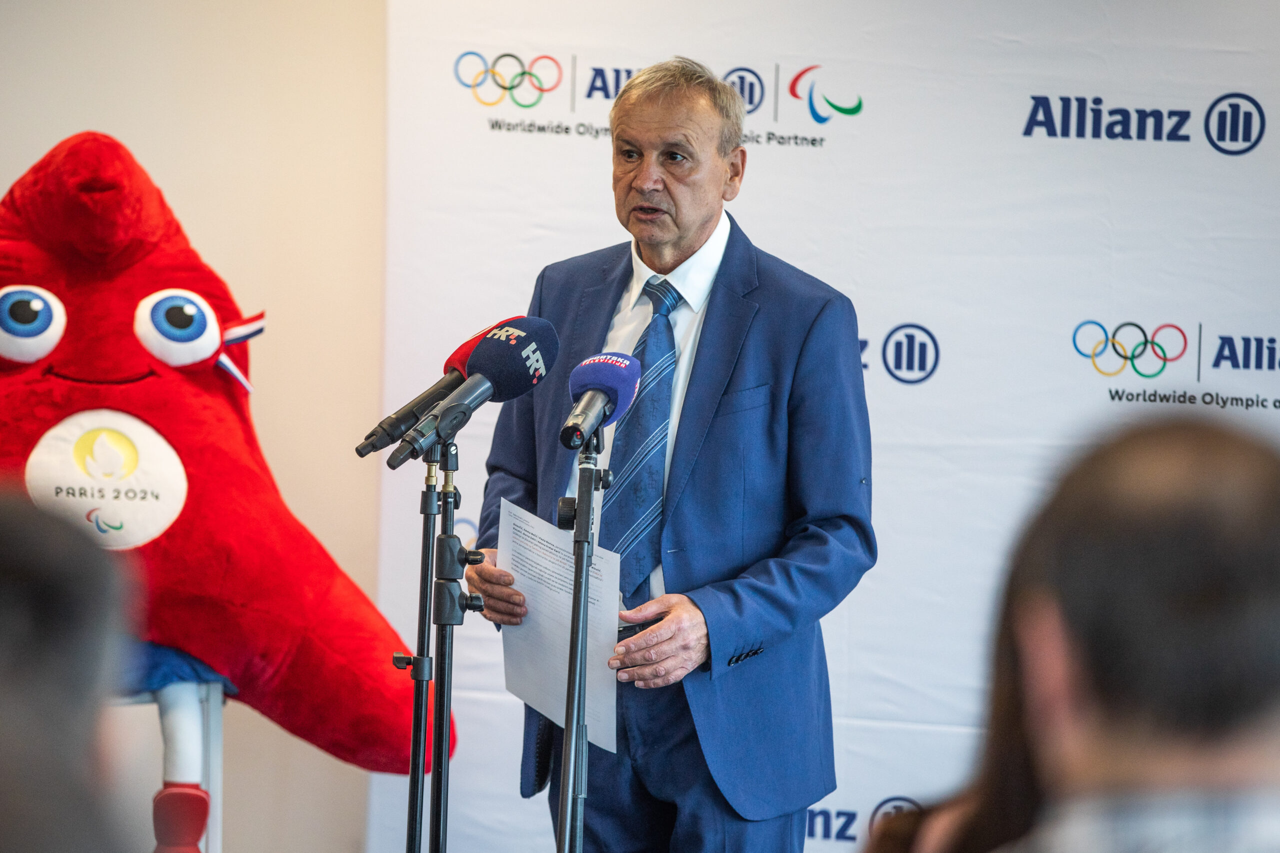 Na slici je čovjek sijede kose u plavom sakou ispred mikrofona, vjerojatno drži govor. Pored njega je crvena maskota, a ispred njega su ljudi.