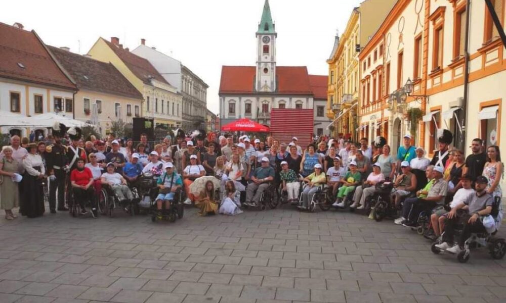 Fotografija prikazuje grupu ljudi koji sjede u kolicima na otvorenom prostoru koji izgleda kao trg u gradu. U pozadini se vide šarene zgrade i crkva s visokim zvonikom. Ova fotografija može biti zanimljiva ili relevantna jer prikazuje događaj ili okupljanje osoba s invaliditetom, što ukazuje na društvenu inkluziju i zajedništvo.