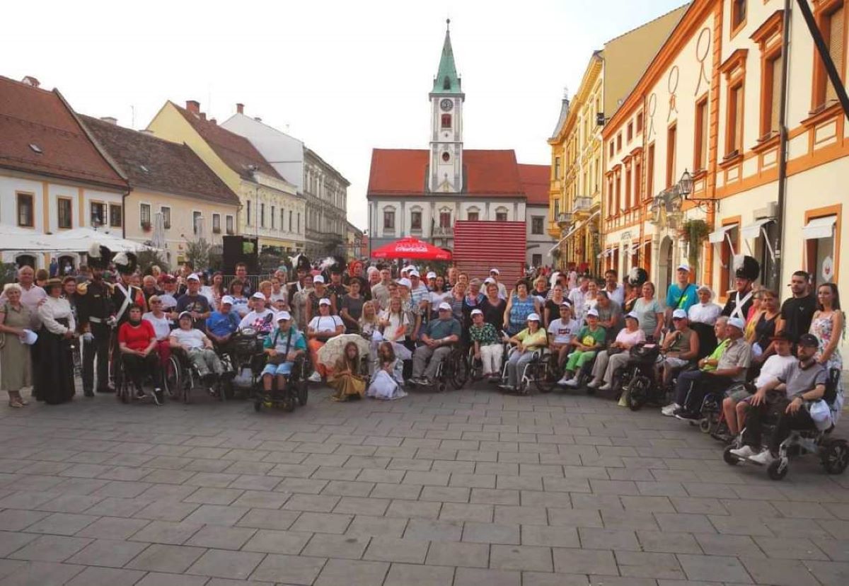 Fotografija prikazuje grupu ljudi koji sjede u kolicima na otvorenom prostoru koji izgleda kao trg u gradu. U pozadini se vide šarene zgrade i crkva s visokim zvonikom. Ova fotografija može biti zanimljiva ili relevantna jer prikazuje događaj ili okupljanje osoba s invaliditetom, što ukazuje na društvenu inkluziju i zajedništvo.