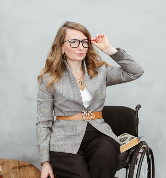 Na slici je žena u invalidskim kolicima. Pored nje je stol na kojem je knjiga. Druga knjiga nalazi se na invalidskim kolicima. Žena je obučena u crne hlače i sivu tanku jaknu sa smeđim remenom i nosi naočale za vid. Jedna ruka joj je spuštena, a drugom popravlja naočale.