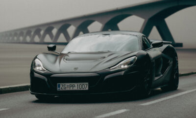 Fotografija prikazuje crni sportski automobil, marke Rimac, parkiran na cesti s mostom u pozadini. Automobil ima zagrebačke registarske tablice.