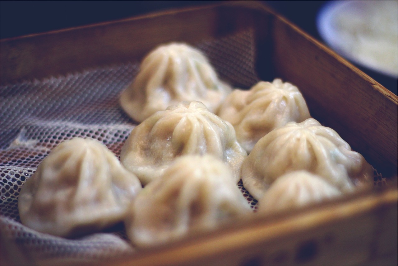 Fotografija prikazuje nekoliko kineskih knedli, poznatih kao dim sum, posluženih u bambusovoj pari. Knedle su bijele boje s naborima na vrhu i izgledaju kao da su upravo skuhane ili pripremljene za posluživanje. Pozadina je zamagljena, što naglašava knedle u prvom planu. Fotografija bi mogla biti zanimljiva zbog detalja i teksture knedli koje sugeriraju svježinu i ukusnost jela, a također može biti relevantna za teme povezane s kineskom kuhinjom ili gastronomijom općenito.