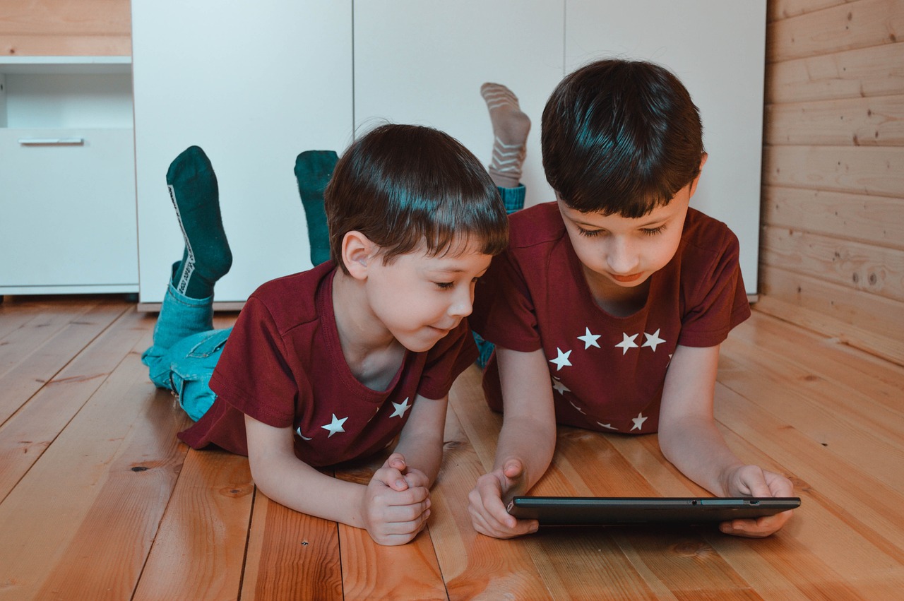 Na fotografiji su dvoje djece u crvenim majicama s bijelim zvijezdama, leže na drvenom podu i gledaju u tablet. Lica djece su zamagljena radi zaštite privatnosti. Zanimljivost ove fotografije može biti moderna tehnologija koja je postala dio svakodnevnog života djece.