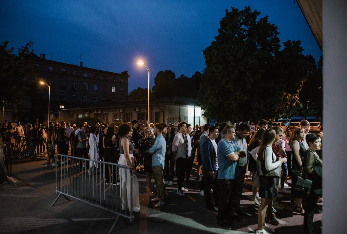 Velika skupina ljudi stoji vani, u redu ispred zgrade. Radi se o večernjem događaju jer je nebo tamno, a ulice osvjetljavaju ulične lampe. Ljudi su obučeni za različite prigode, a atmosfera je društvena i živahna.