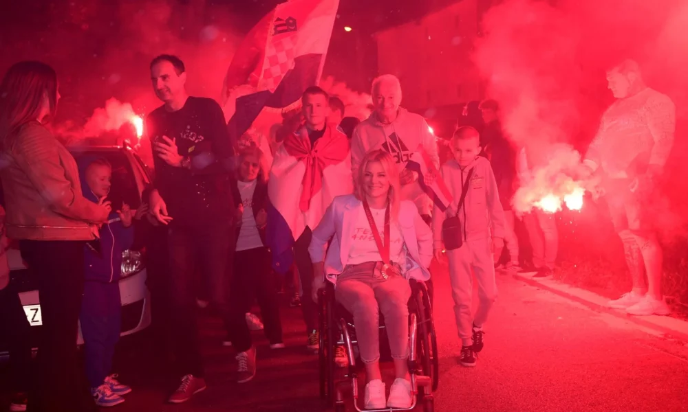 Fotografija prikazuje više ljudi i zapaljene baklje u pozadini. Jedna ženska osoba sjedi u kolicima, a oko vrata ima medalju. Oko nje je više veselih ljudi, a iza nje nalazi se i čovjek zaogrnjen hrvatskim barjakom dok drugi hrvatski barjak visi viori u zraku.