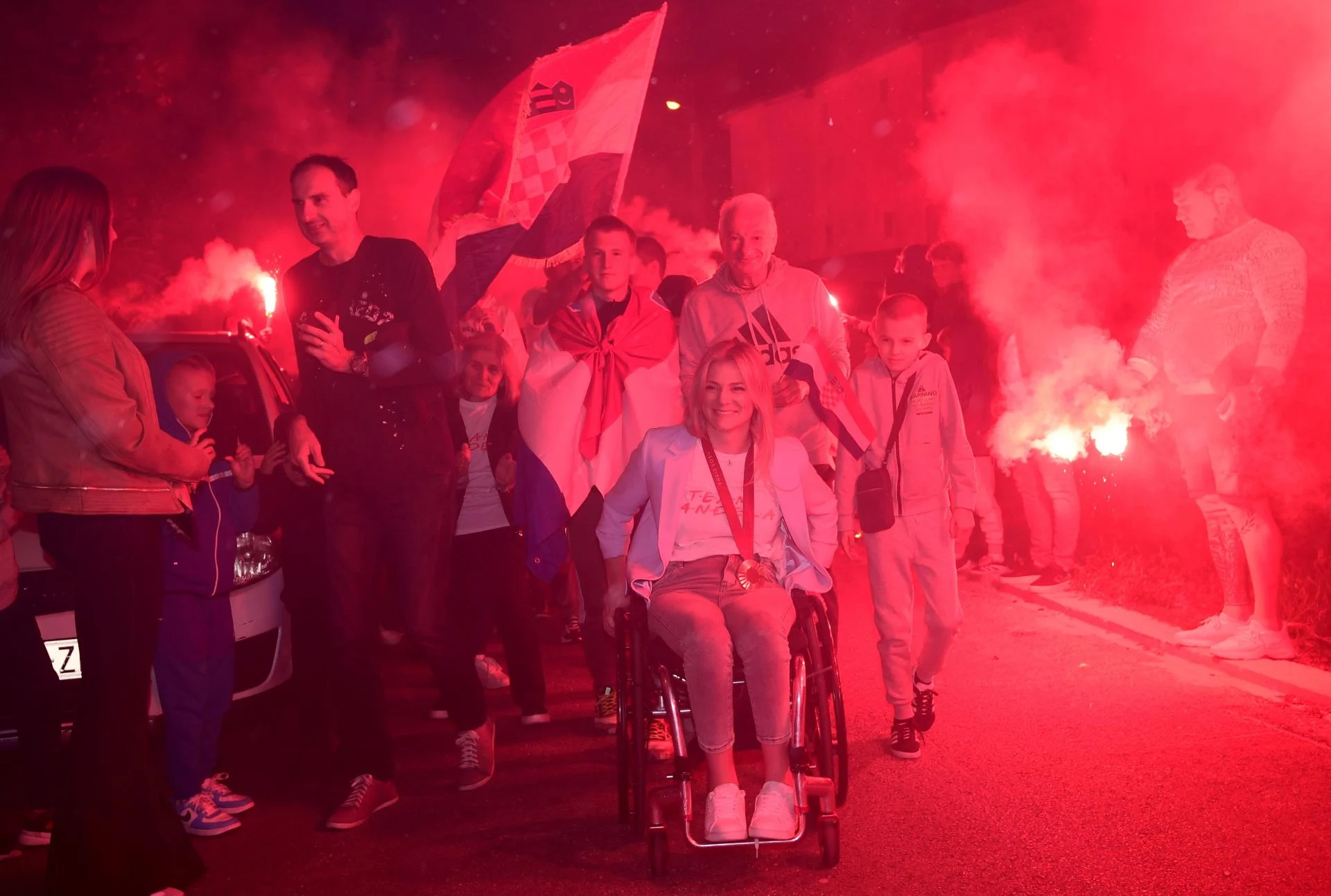 Fotografija prikazuje više ljudi i zapaljene baklje u pozadini. Jedna ženska osoba sjedi u kolicima, a oko vrata ima medalju. Oko nje je više veselih ljudi, a iza nje nalazi se i čovjek zaogrnjen hrvatskim barjakom dok drugi hrvatski barjak visi viori u zraku.