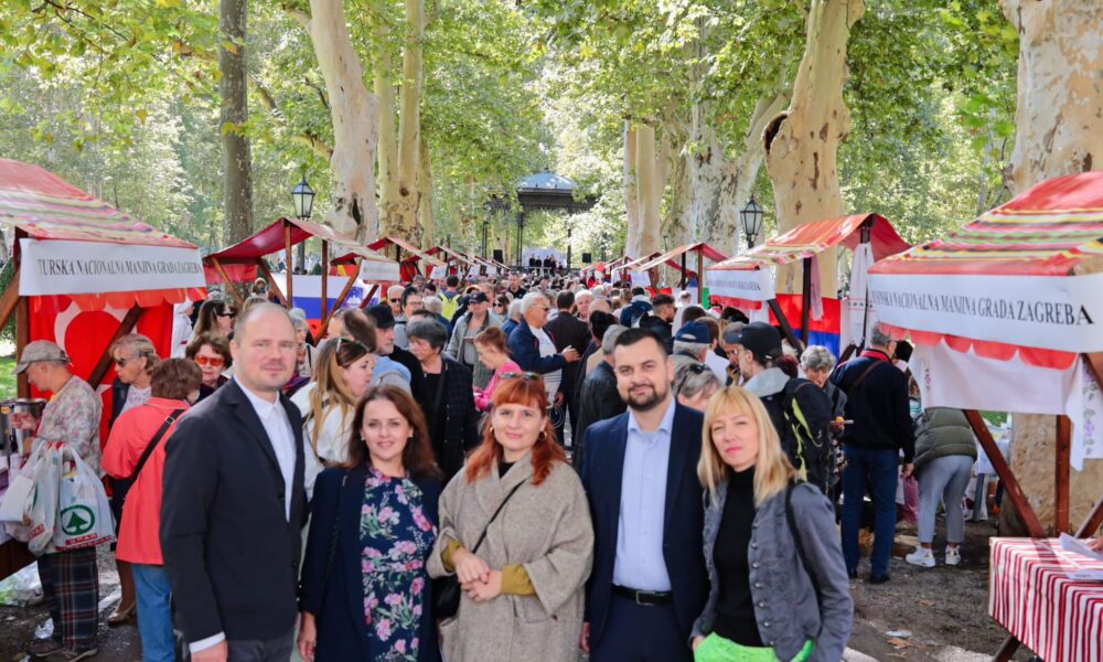 Slika prikazuje događaj na otvorenom pod nazivom "Dan nacionalnih manjina". Okruženi drvećem, štandovi različitih nacionalnih manjina Grada Zagreba postavljeni su uzduž šetnice. Na štandovima se vide razni predmeti i natpisi, a u središtu slike pozira pet osoba, od kojih su četiri žene i jedan muškarac. U pozadini se vidi mnoštvo posjetitelja koji šetaju i razgledavaju štandove. Atmosfera je vesela, a događaj se odvija u prirodnom okruženju s puno zelenila.