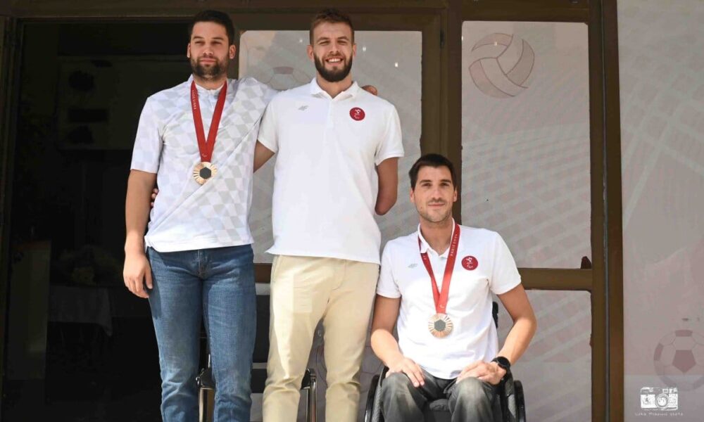 Na fotografiji je prikazano troje ljudi od kojih dvoje nosi brončane medalje oko vrata. Jedna osoba od tih troje je u invalidskim kolicima. Svi nose bijele majice i različite hlače. S obzirom na to da imaju medalje upućuje na to da su se vratili s nekog natjecanja i ostvarili sjajne rezultate.