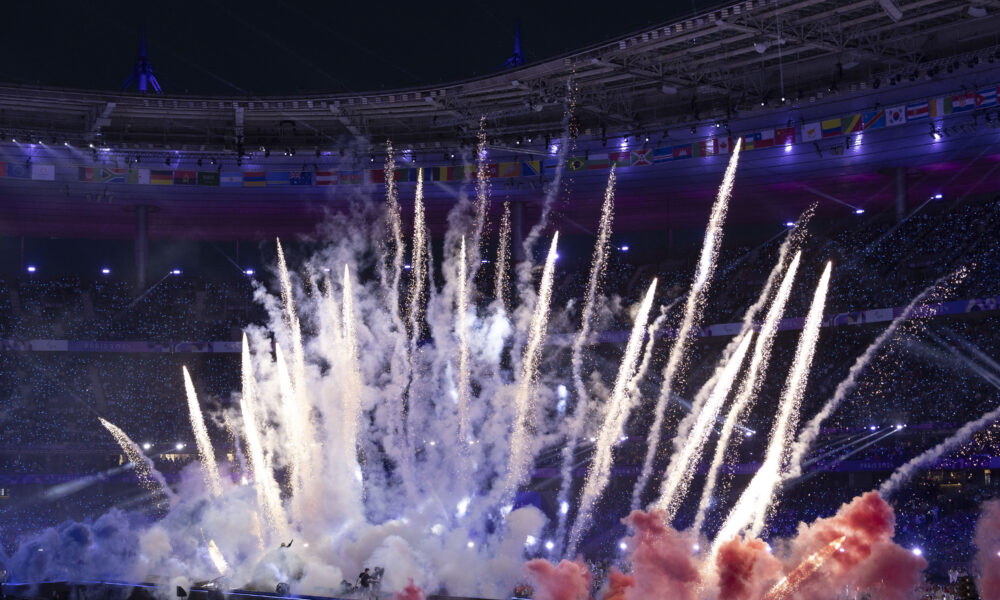 Slika prikazuje vatromet unutar velikog stadiona noću, s više mlazova svjetla i dima koji se dižu u zrak. Na tribinama u pozadini vide se mnoštvo zastava različitih zemalja, dok su tribine pune publike. Stadion je osvijetljen plavim i ljubičastim svjetlima, stvarajući dramatičnu atmosferu. Na tlu ispod vatrometa vidi se pozornica s izvođačima u daljini, okruženima dimom u raznim bojama, uključujući crvenu i plavu.