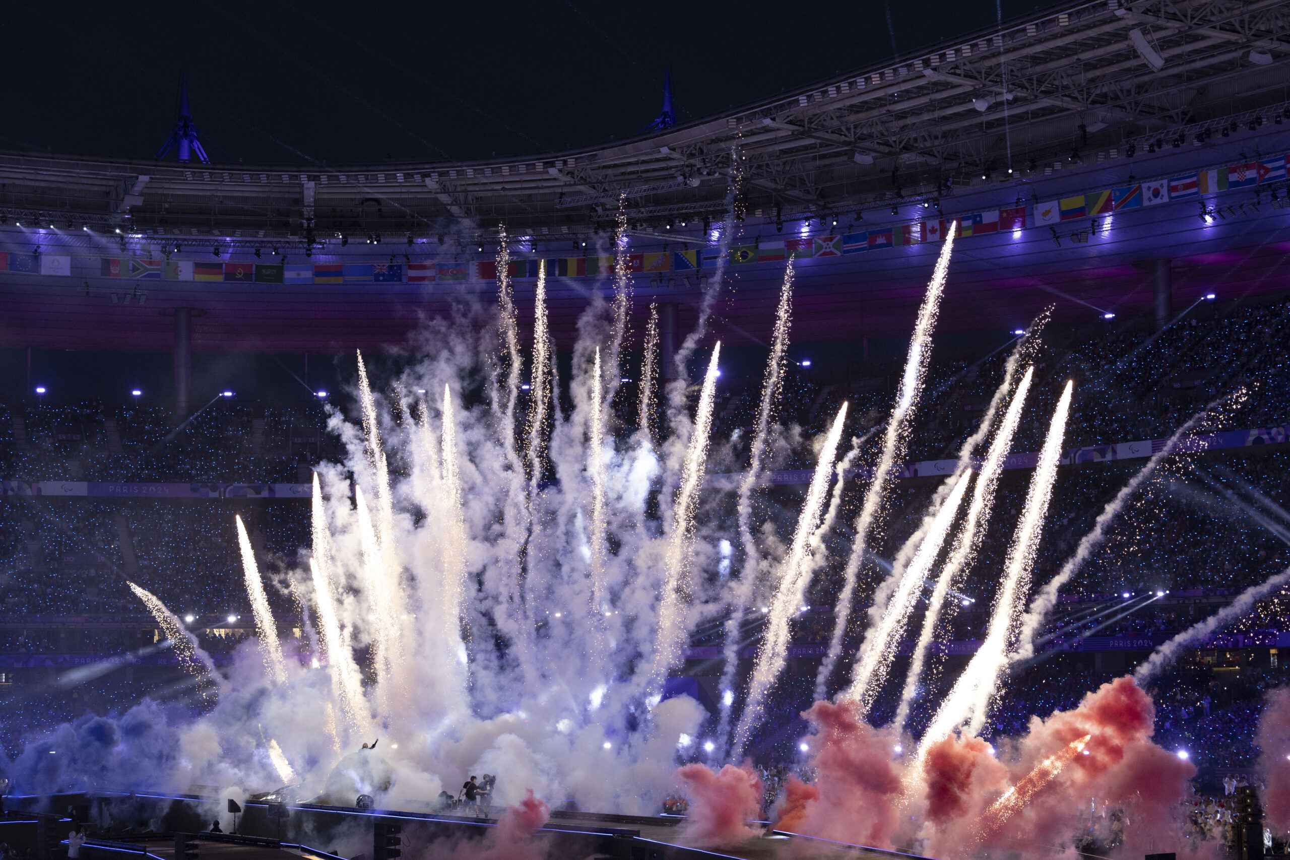 Slika prikazuje vatromet unutar velikog stadiona noću, s više mlazova svjetla i dima koji se dižu u zrak. Na tribinama u pozadini vide se mnoštvo zastava različitih zemalja, dok su tribine pune publike. Stadion je osvijetljen plavim i ljubičastim svjetlima, stvarajući dramatičnu atmosferu. Na tlu ispod vatrometa vidi se pozornica s izvođačima u daljini, okruženima dimom u raznim bojama, uključujući crvenu i plavu.