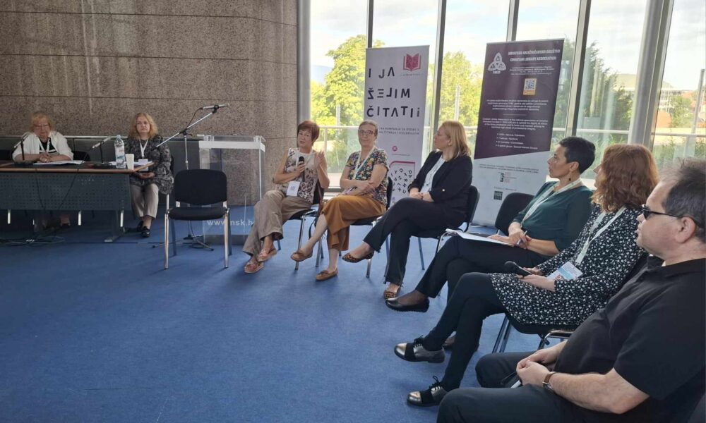 Na fotografiji je prikazano nekoliko ljudi koji sjede u krug i nešto raspravljaju. Vjerojatno na nekoj konferenciji. Iza njih su dva panoa na kojima nešto piše. Osim ljudi koji sjede na stolicama, na fotografiji su prikazane i dvije žene koje sjede za crnim stolom. Konferencija se održala u ugodnom i osvjetljenom unutarnjem prostoru.