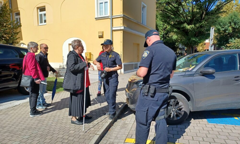 Na slici su dva policajca u uniformama koja razgovaraju sa starijom ženom koja koristi bijeli štap za slijepe osobe. Iza nje su još dvije osobe, jedna žena i jedan muškarac, koji također drže štapove za hodanje ili kišobrane. Scena se odvija ispred zgrade s žutim zidom i parkiranim automobilom, a policajci djeluju angažirano u interakciji s prisutnima. Atmosfera je mirna i čini se da se odvija neka vrsta informativnog razgovora ili provjere.